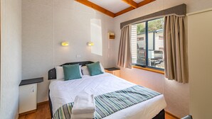 Una cama bien hecha con colcha a rayas, una almohada blanca y una toalla doblada. Hay una mesita de noche con una lámpara y una ventana con cortinas.