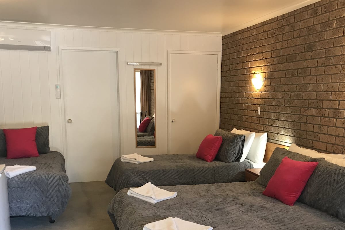 A hotel room with two beds, each with grey bedding and red pillows, a white door, a mirror, and a brick wall with a wall-mounted light.