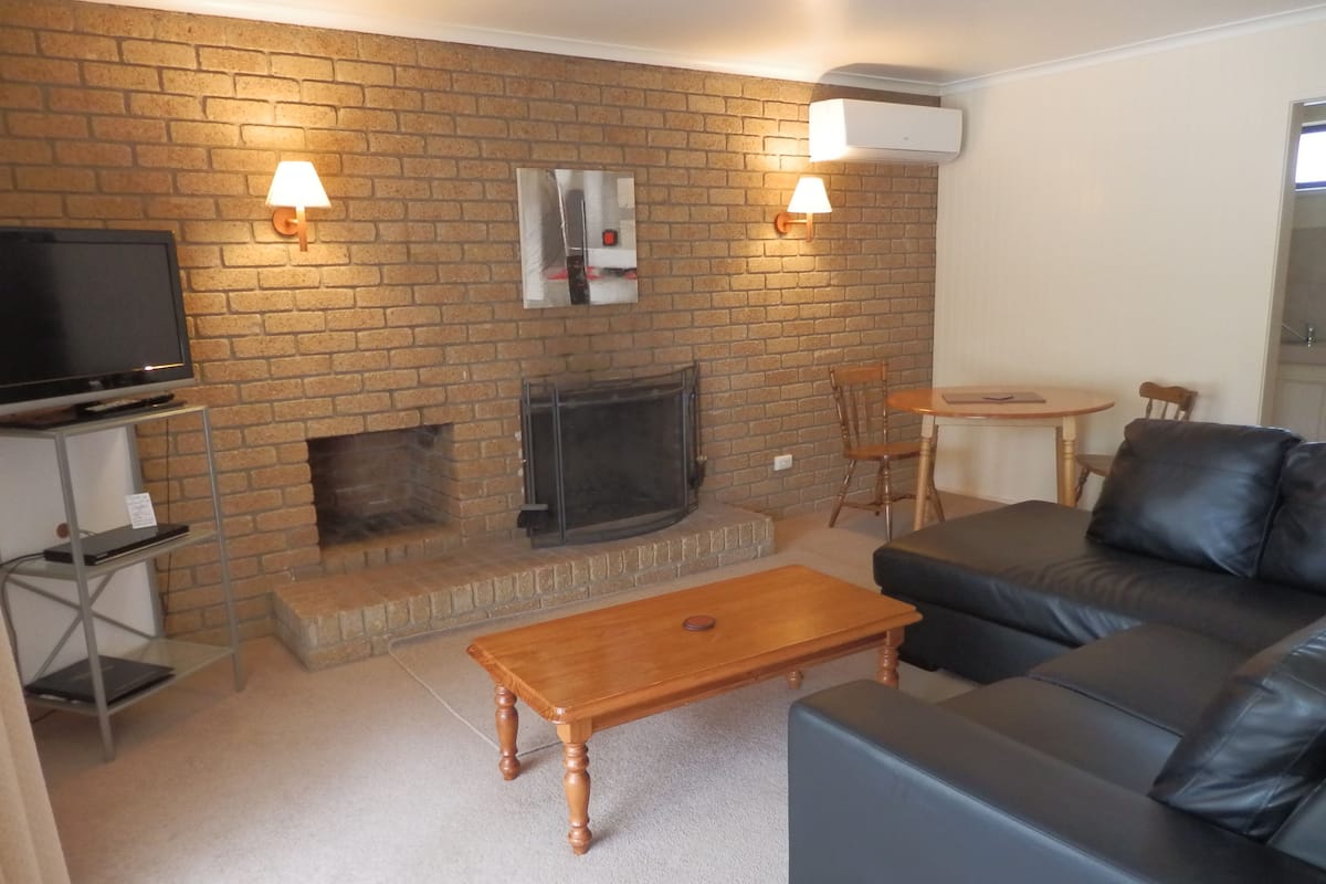 A living room with a brick fireplace, a television on a stand, a leather sofa, and two wall-mounted lamps.