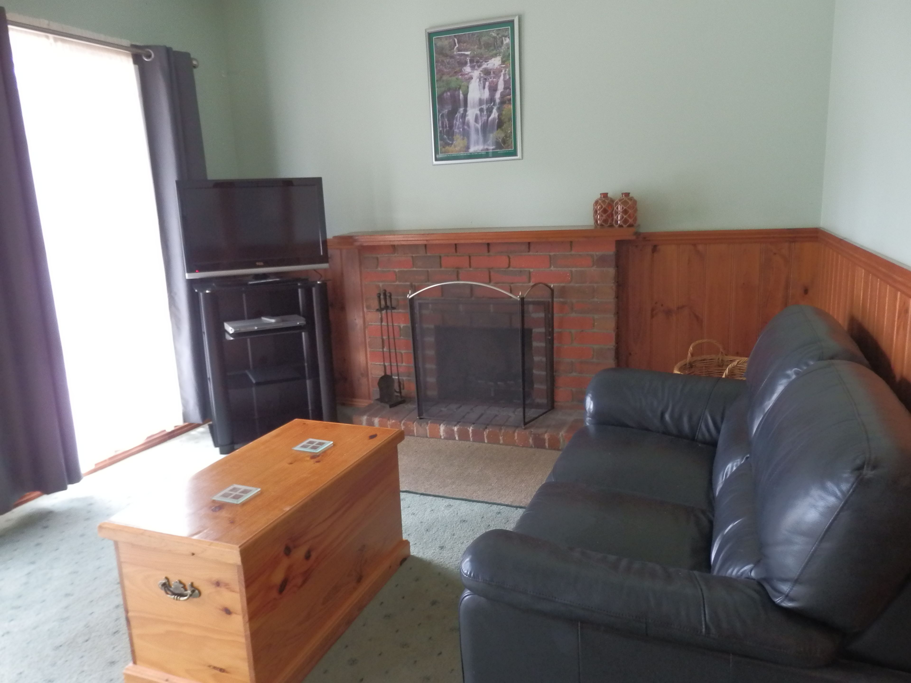 Una sala de estar con chimenea, un sofá de cuero negro, un cofre de madera y un televisor.