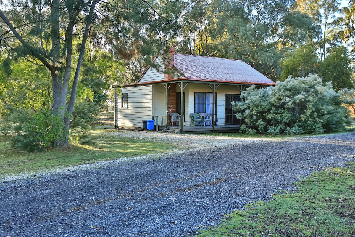 A single-story house with a porch, surrounded by trees and a gravel driveway.