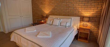 A bedroom with a brick wall, a bed with white bedding, two bedside tables with lamps, and a wardrobe.