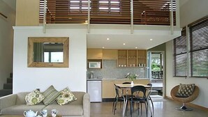 A modern living room with a sofa, a dining table with chairs, a kitchen area, and a balcony with wooden blinds. - Elfin Hill Country Accommodation (Pokolbin)