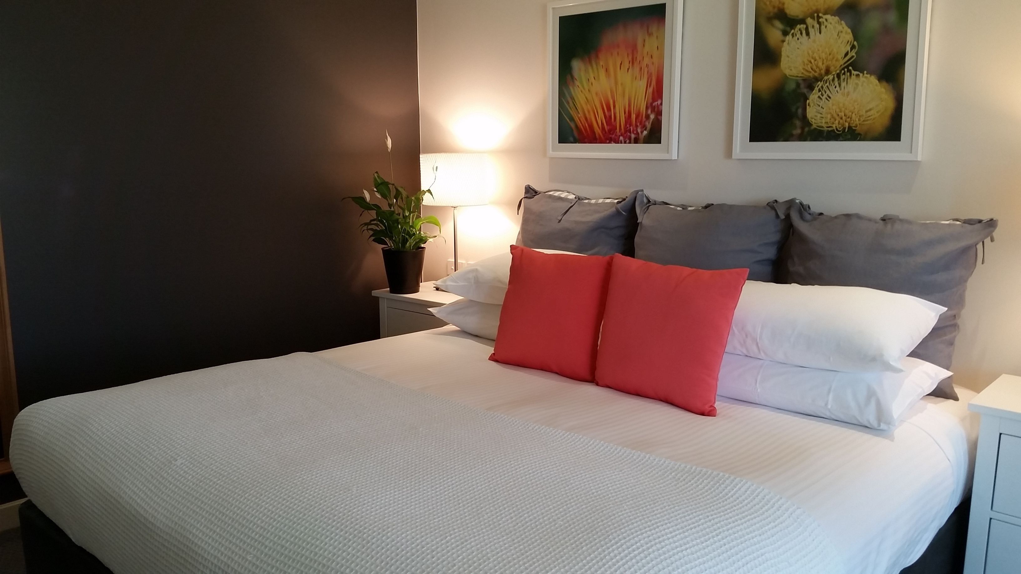A neatly made bed with white and red pillows, a bedside lamp, and two framed floral pictures on the wall.