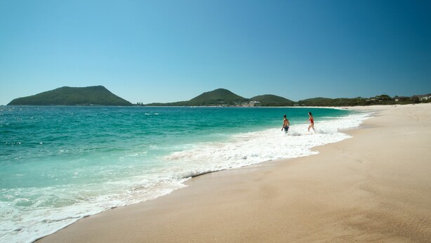 A beach with clear turquoise water, a sandy shore, and a mountain in the background. - Bali at the Bay (Nelson Bay)