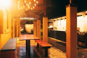 Un patio al aire libre con mesa y bancos de madera, una zona de cocina con estufa y fregadero, y un paseo techado con guirnaldas de luces.