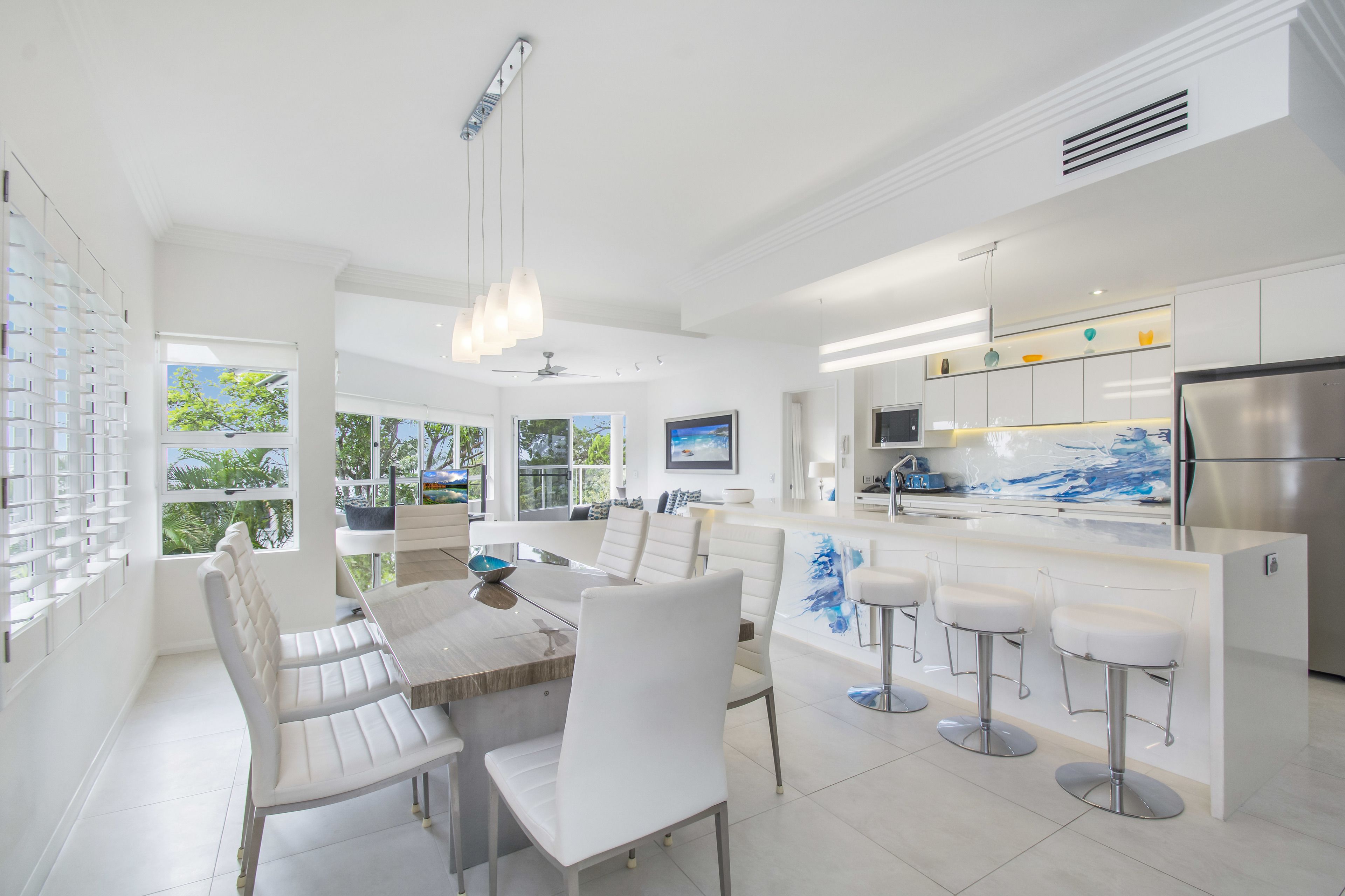 Una cocina moderna con armarios blancos, una isla central con taburetes y un comedor con mesa de madera y sillas blancas.