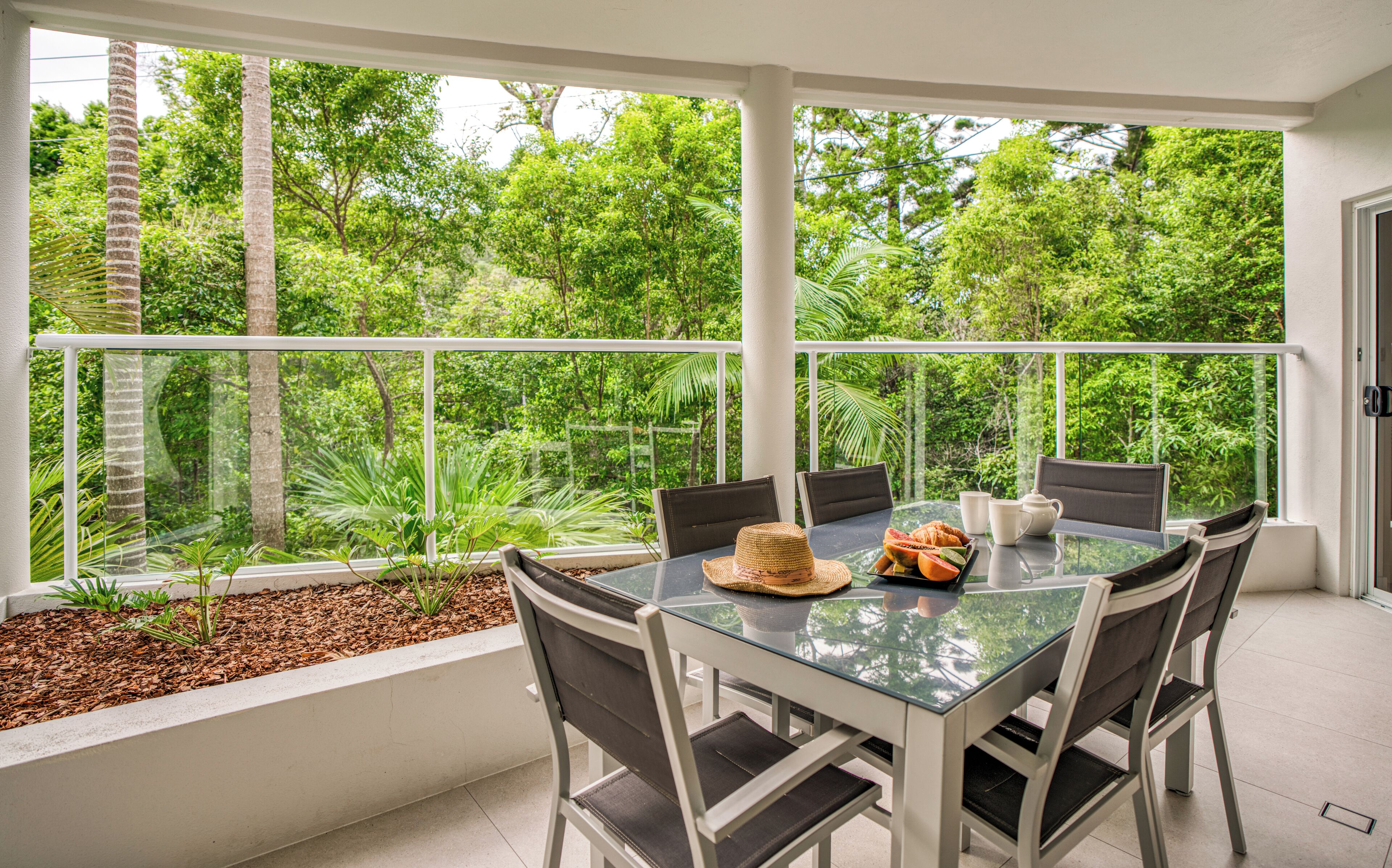 A glass-top table with chairs, a woven hat, a fruit bowl, and a teapot on a balcony with a view of lush greenery.