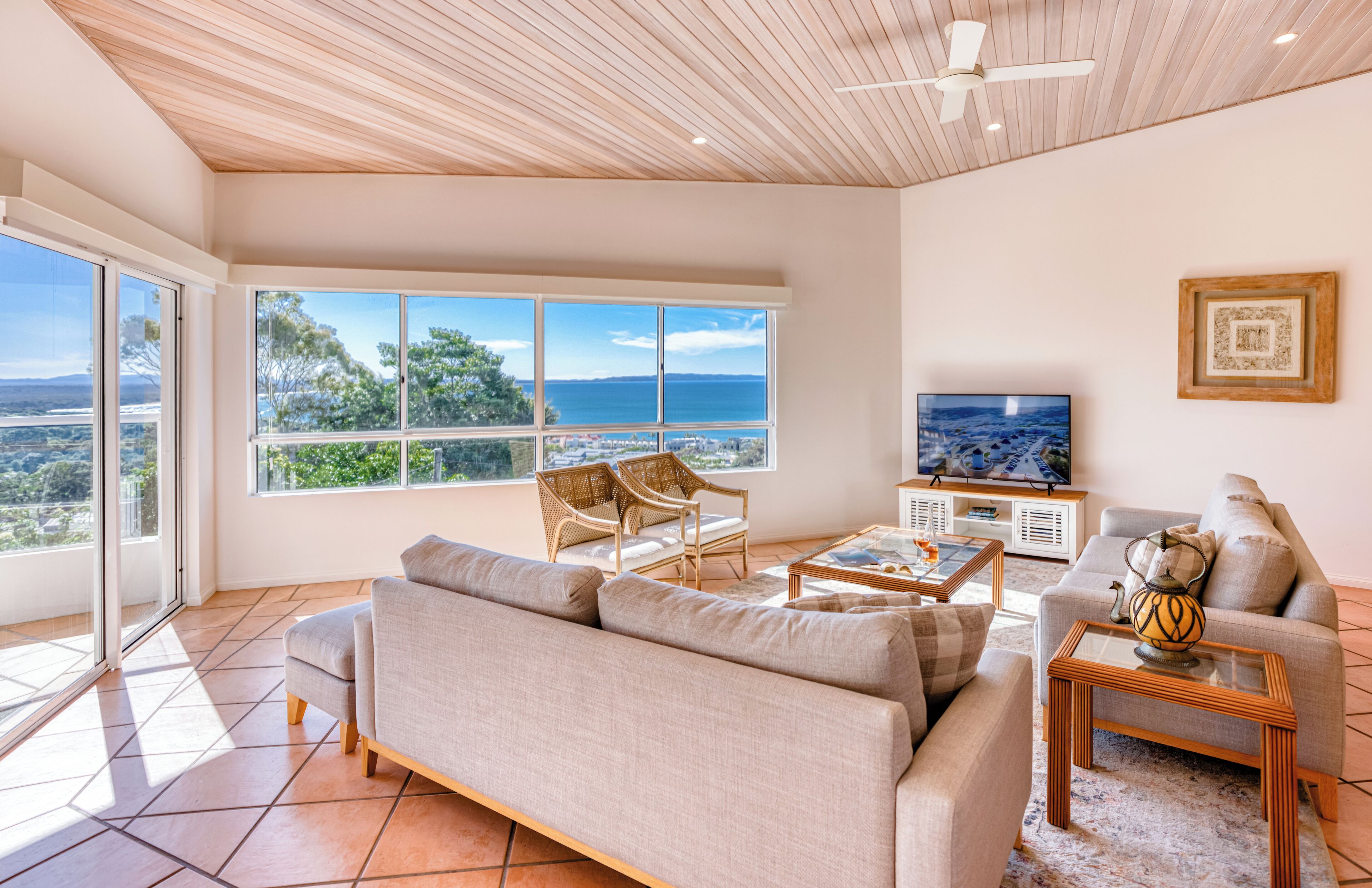 A spacious living room with a large sofa, two armchairs, a coffee table, and a TV. The room has large windows offering a view of the sea and a cityscape.