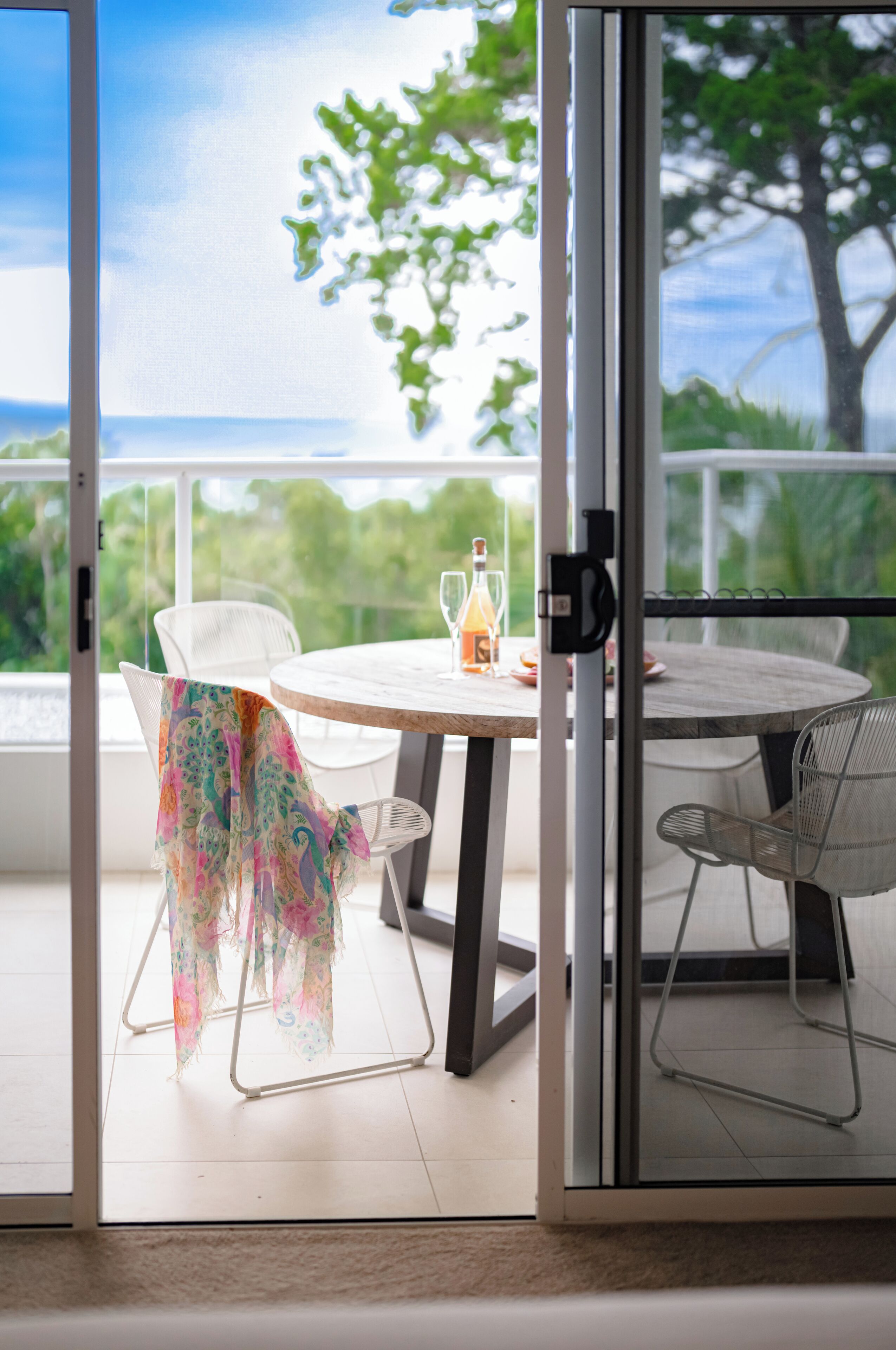 A room with a glass door leading to a balcony with a table and chairs, a bottle of drink, and a view of greenery.