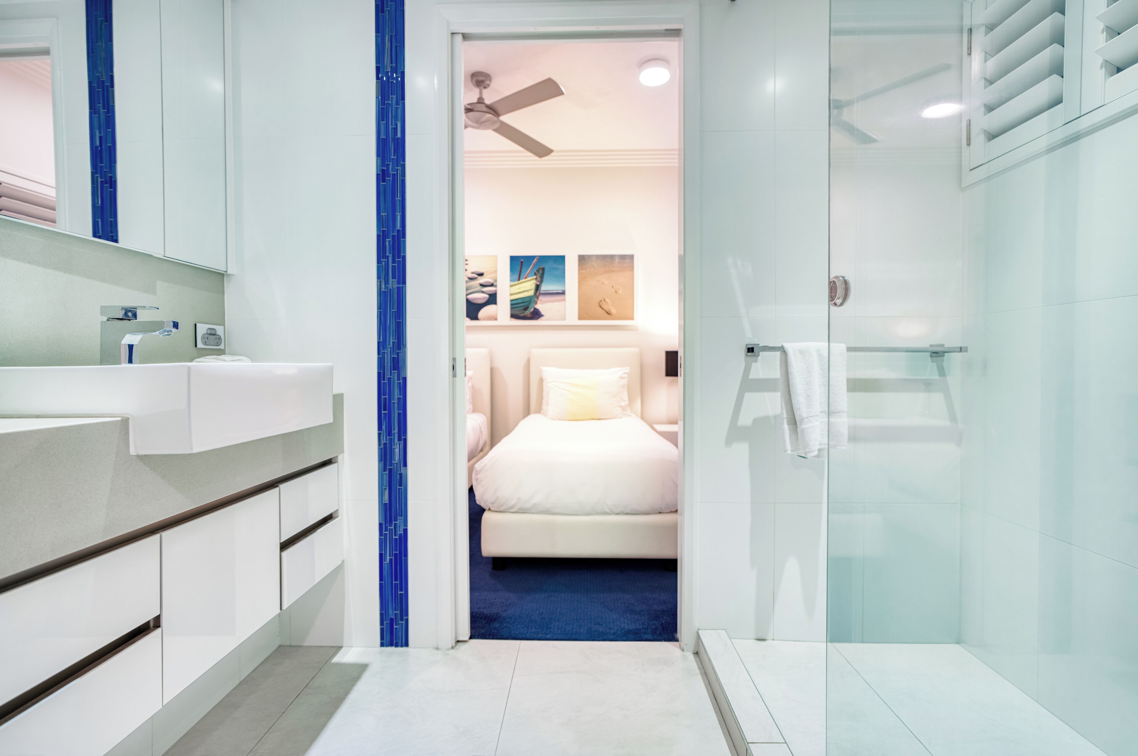 A modern bathroom with a glass shower enclosure, a white sink, and a bed visible through an open door.