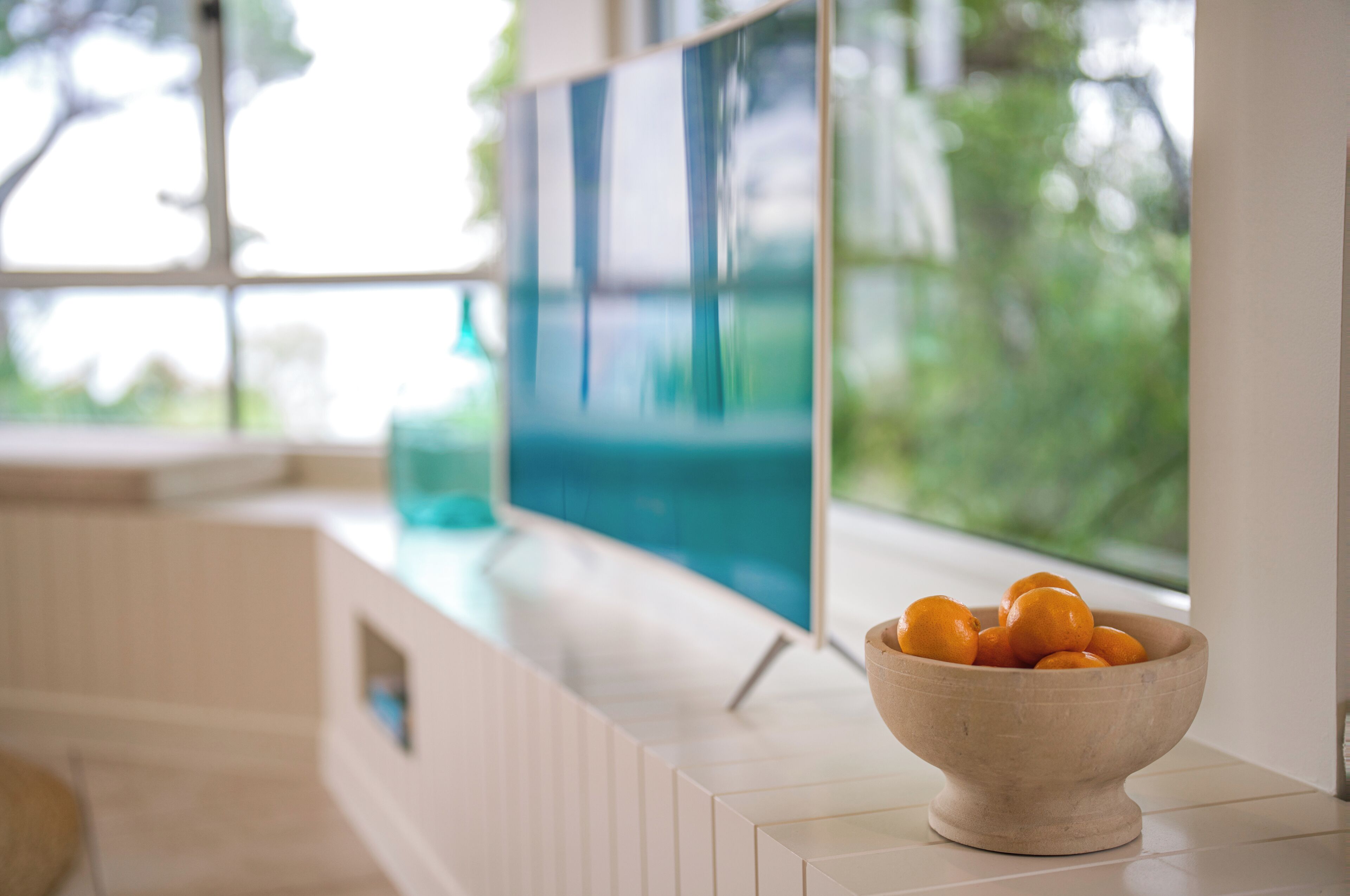 Une cuisine moderne avec une grande fenêtre, un saladier en bois rempli d’oranges posé sur le comptoir, et un téléviseur à écran plat fixé au mur.