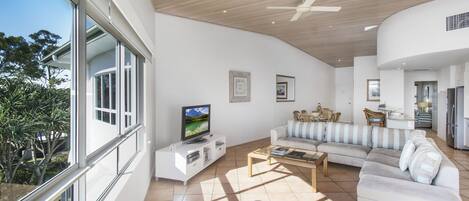 A spacious living area with a large window, a flat-screen TV, a sofa, a coffee table, and a ceiling fan.