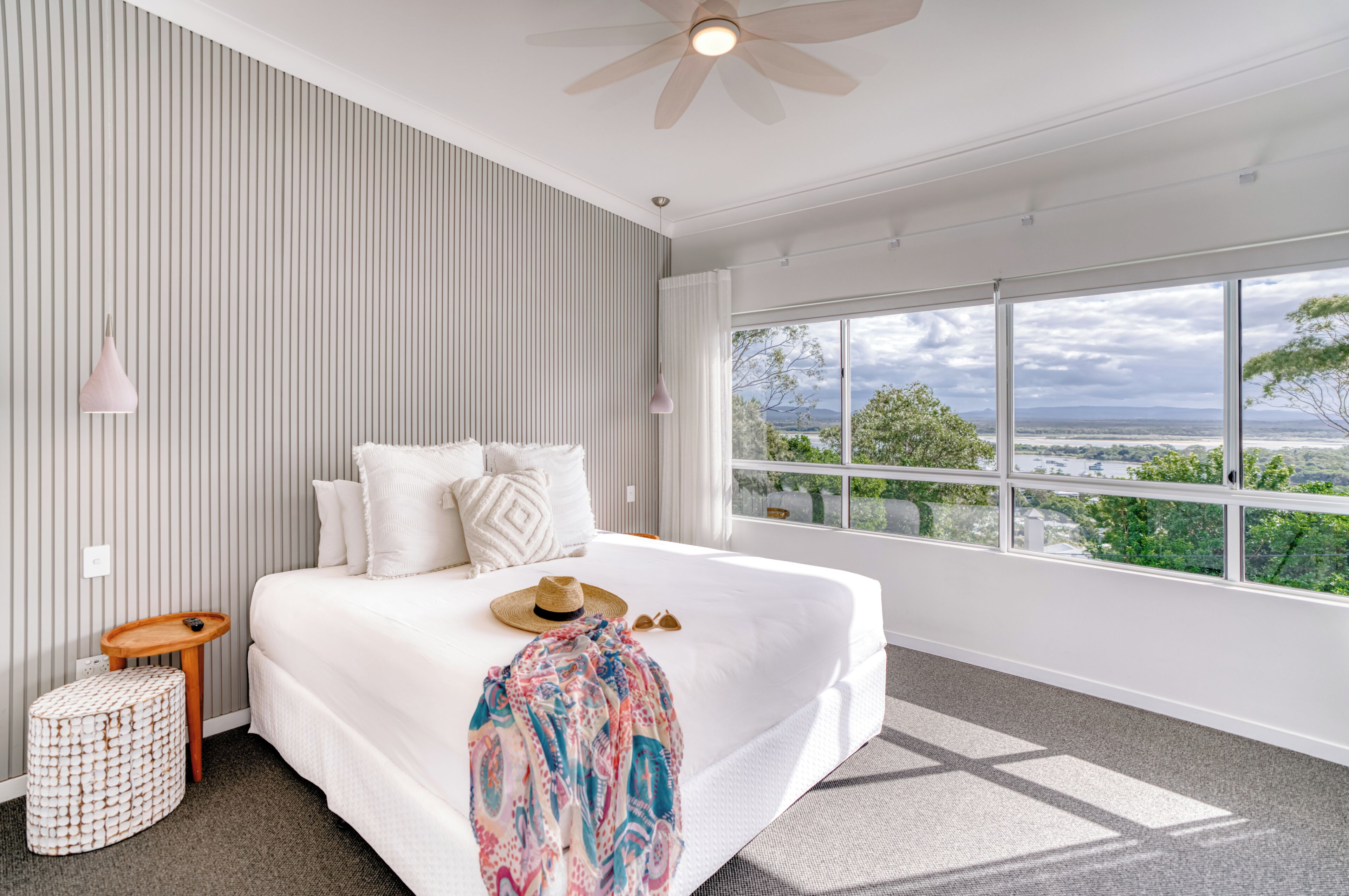 A bedroom with a large bed, a ceiling fan, a view of the outdoors, and a window with curtains.