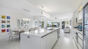 A modern kitchen with white cabinetry, a central island, and a dining area with a white table and chairs. - The Lookout Resort (Noosa Heads)