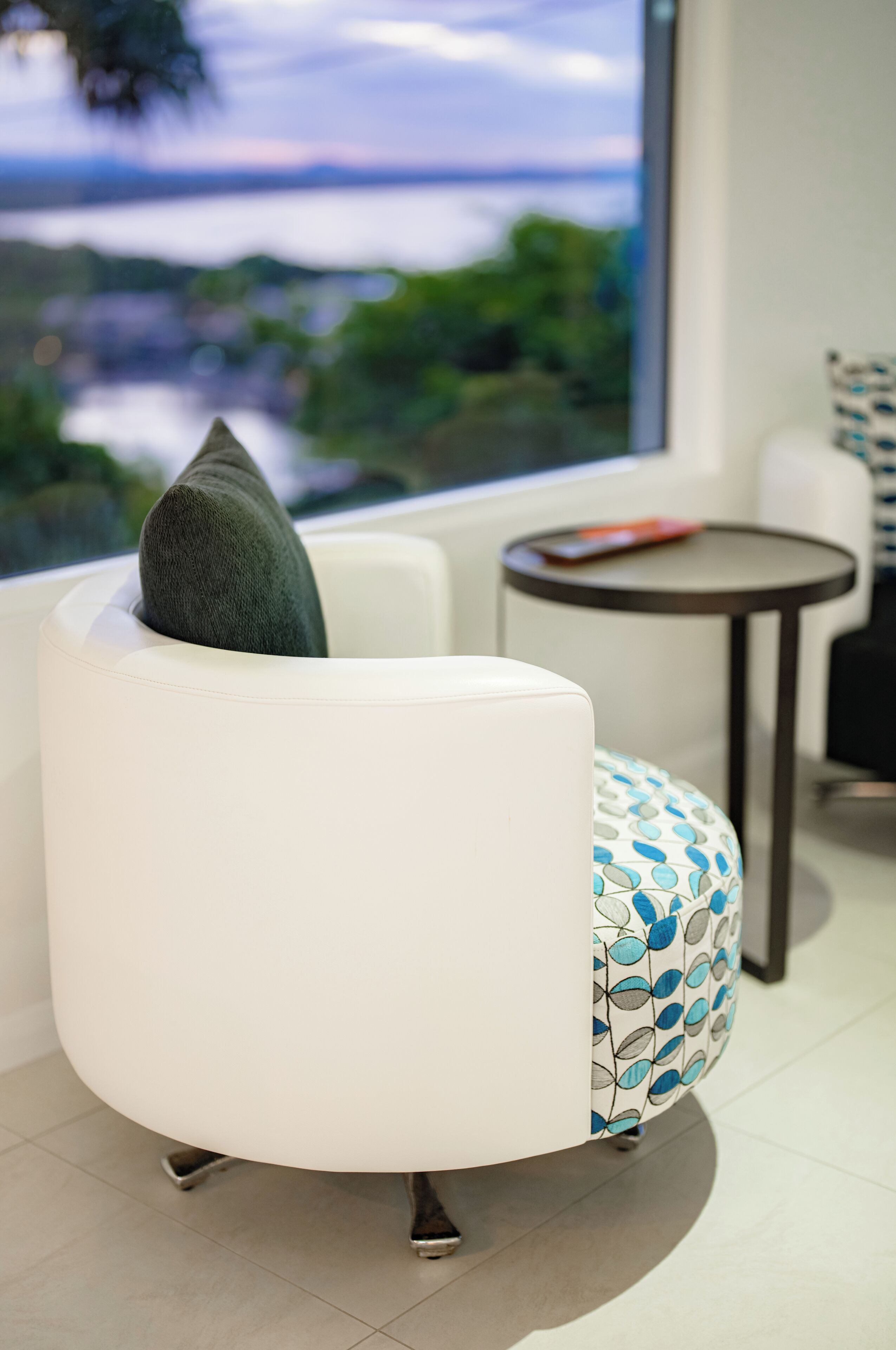 Una sala de estar moderna con un sofá blanco, un otomano estampado y vista a un cuerpo de agua a través de un ventanal grande.