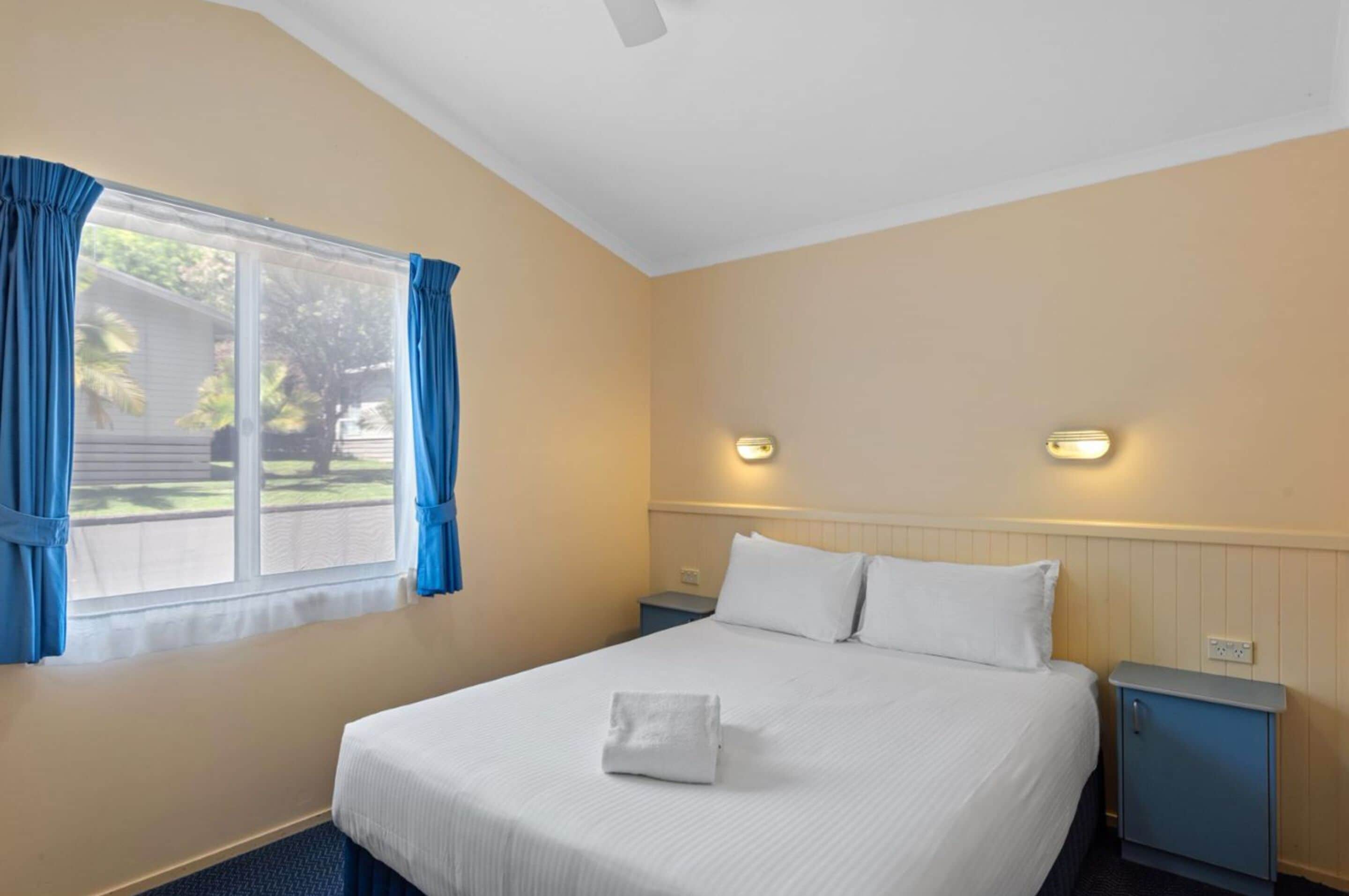 Een hotelkamer met een bed, nachtkastje, wandlamp en een raam met gordijnen.