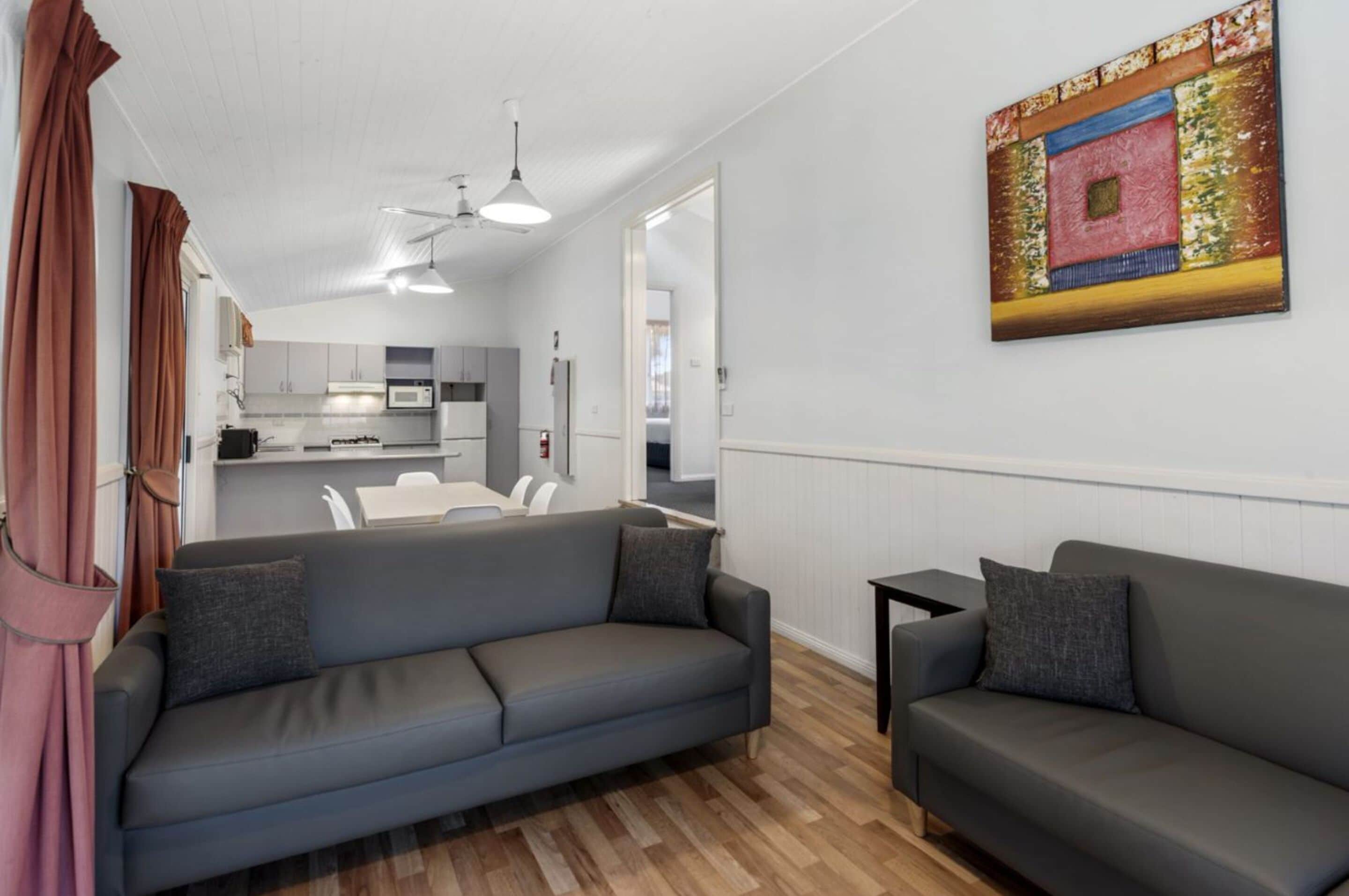 A modern living room with two grey sofas, a wooden floor, a kitchen in the background, and a painting on the wall.