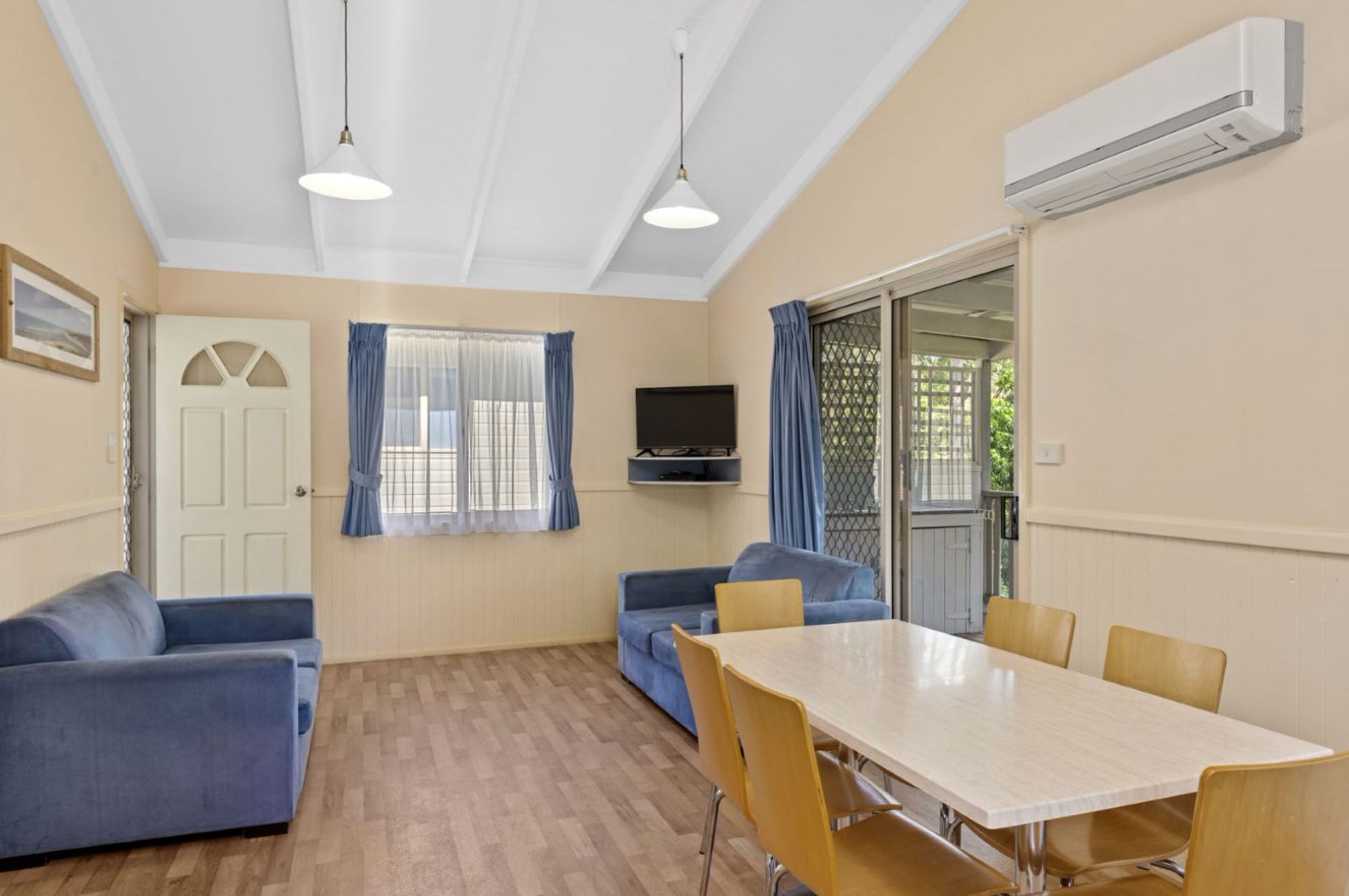 A room with a wooden floor, two blue sofas, a white table with chairs, a TV, and an air conditioning unit.