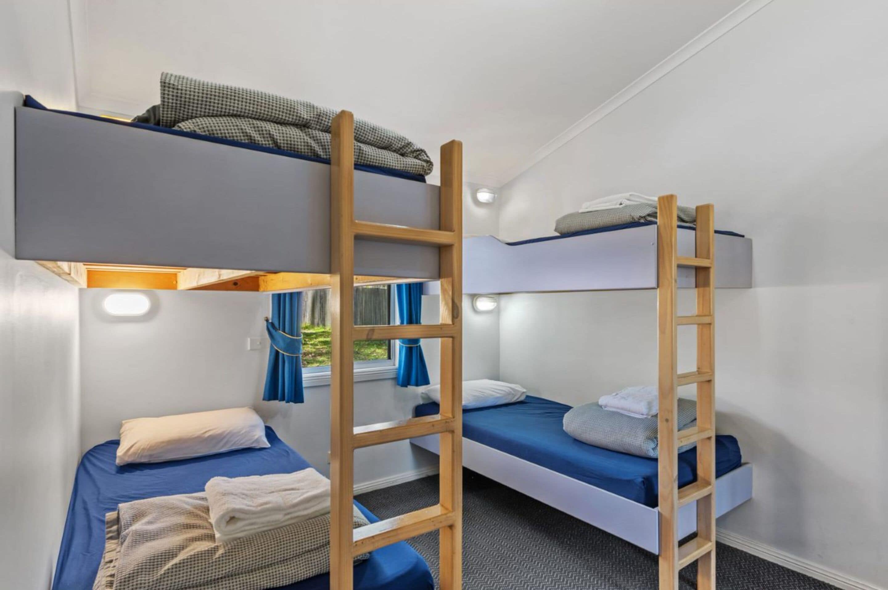 Une chambre avec un lit superposé, des échelles en bois, des murs blancs et une fenêtre avec des rideaux bleus.