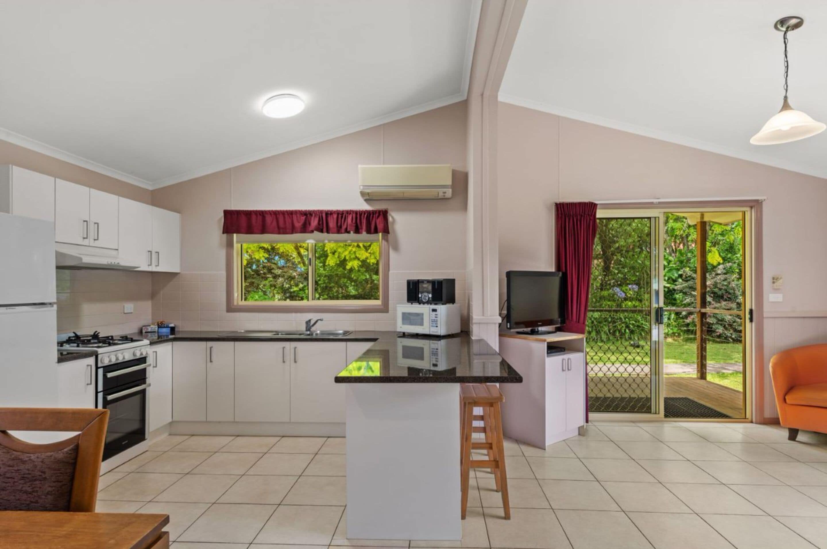 A modern kitchen with white cabinets, a central island, a gas stove, and a microwave. There is a dining area with a table and chairs, and a sliding glass door leading to a balcony with a view of greenery.