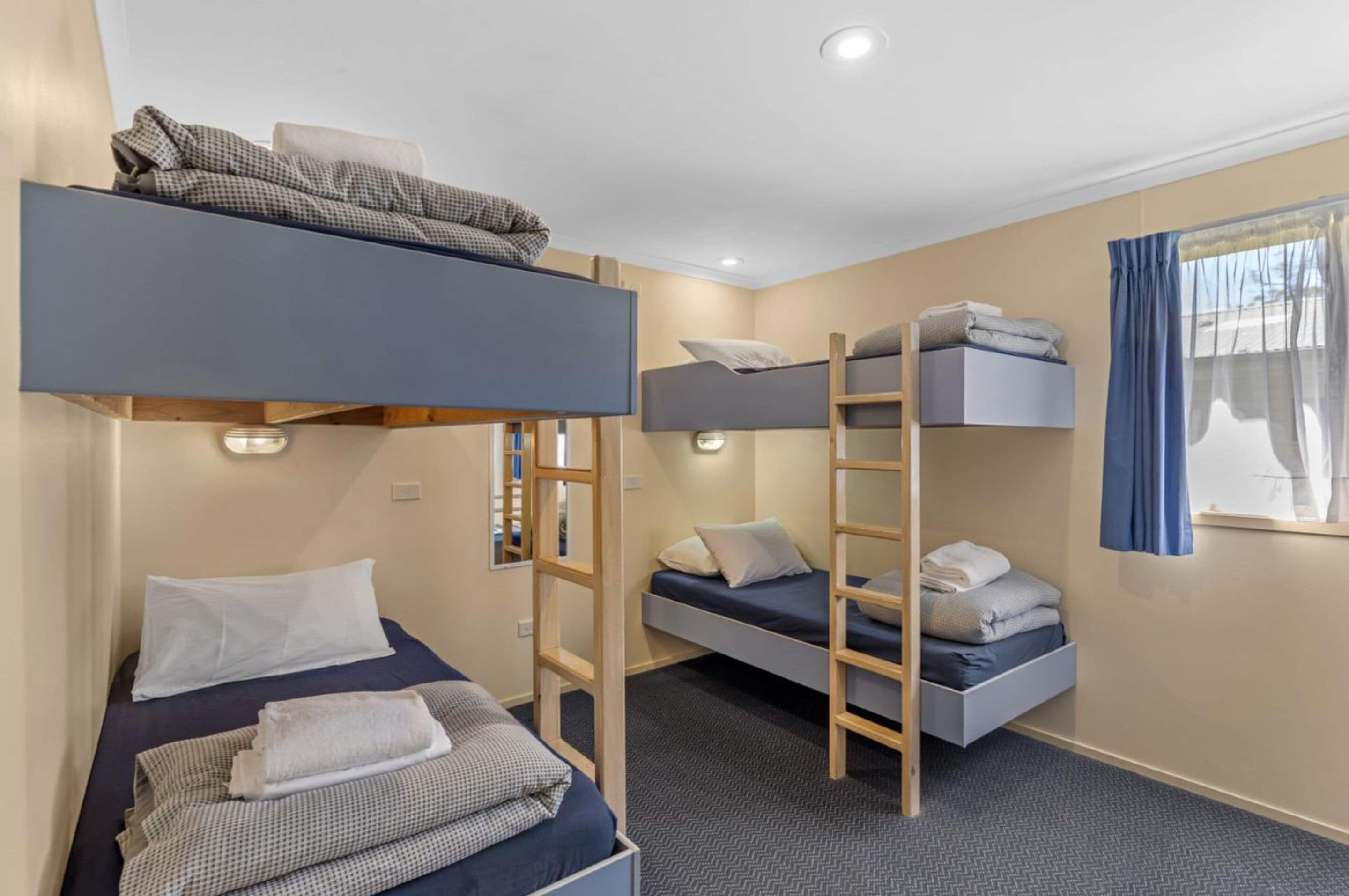 A dormitory room with bunk beds, a ladder, a small desk, and a window with curtains.