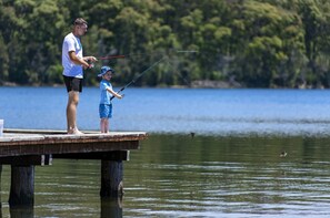 A man and a child fishing on a wooden pier extending into a lake, surrounded by trees. - Discovery Parks - Ulladulla (Burrill Lake)