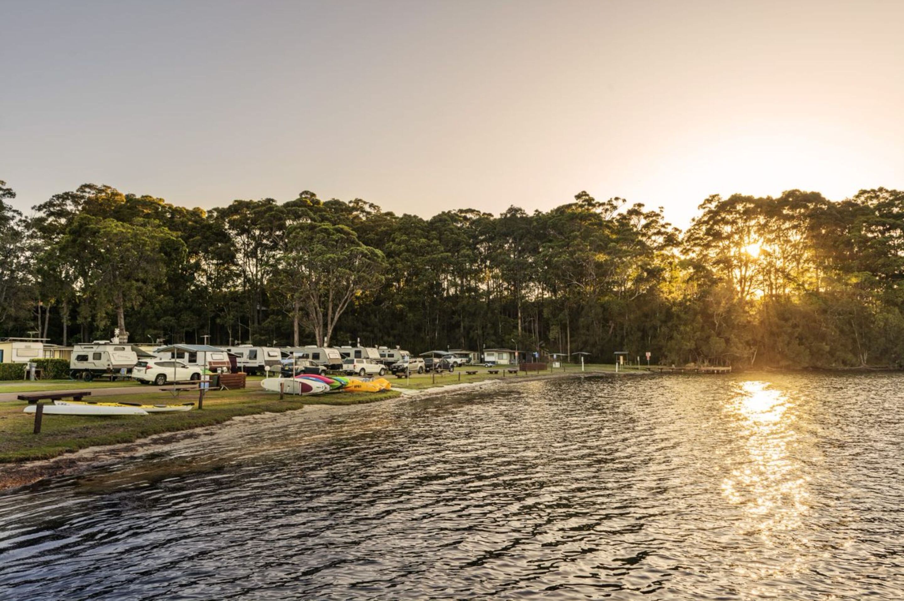 Un campamento junto al lago con vehículos recreativos estacionados a lo largo de la orilla, rodeado de árboles y una vista despejada del atardecer.