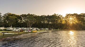 Un campamento junto al lago con vehículos recreativos estacionados a lo largo de la orilla, rodeado de árboles y una vista despejada del atardecer.