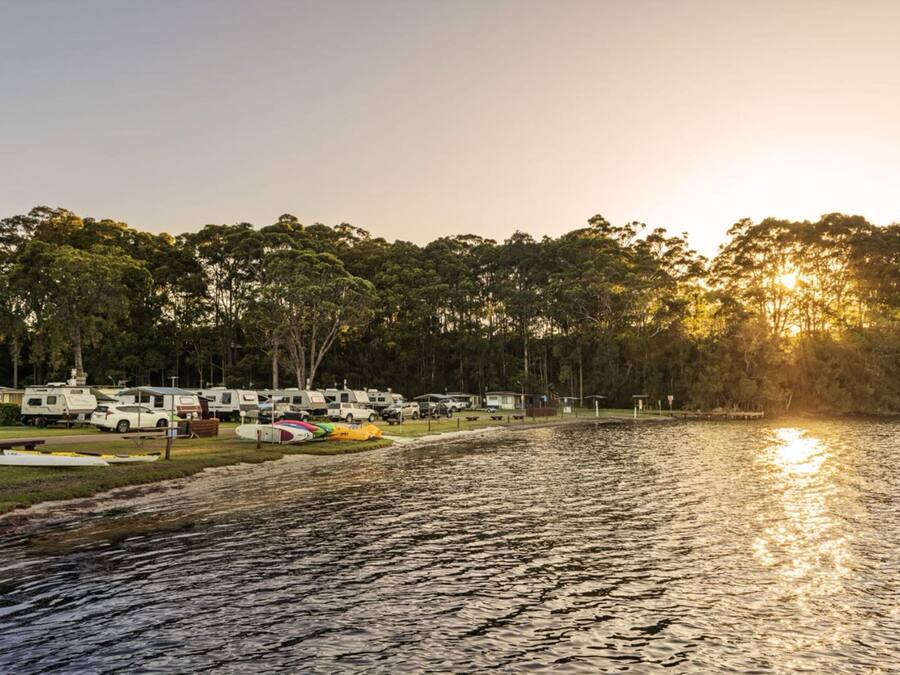 Un campamento junto al lago con vehículos recreativos estacionados a lo largo de la orilla, rodeado de árboles y una vista despejada del atardecer.