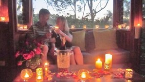 Una pareja sentada a una mesa con velas y una bebida, en un ambiente acogedor con vistas a árboles.