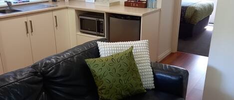 A modern living room with a black leather sofa, a small kitchen area with a microwave and dishwasher, and two wall-mounted paintings.