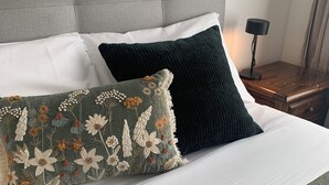 Una cama bien tendida con un cabecero tapizado en gris, ropa de cama blanca y dos almohadones con motivos florales.