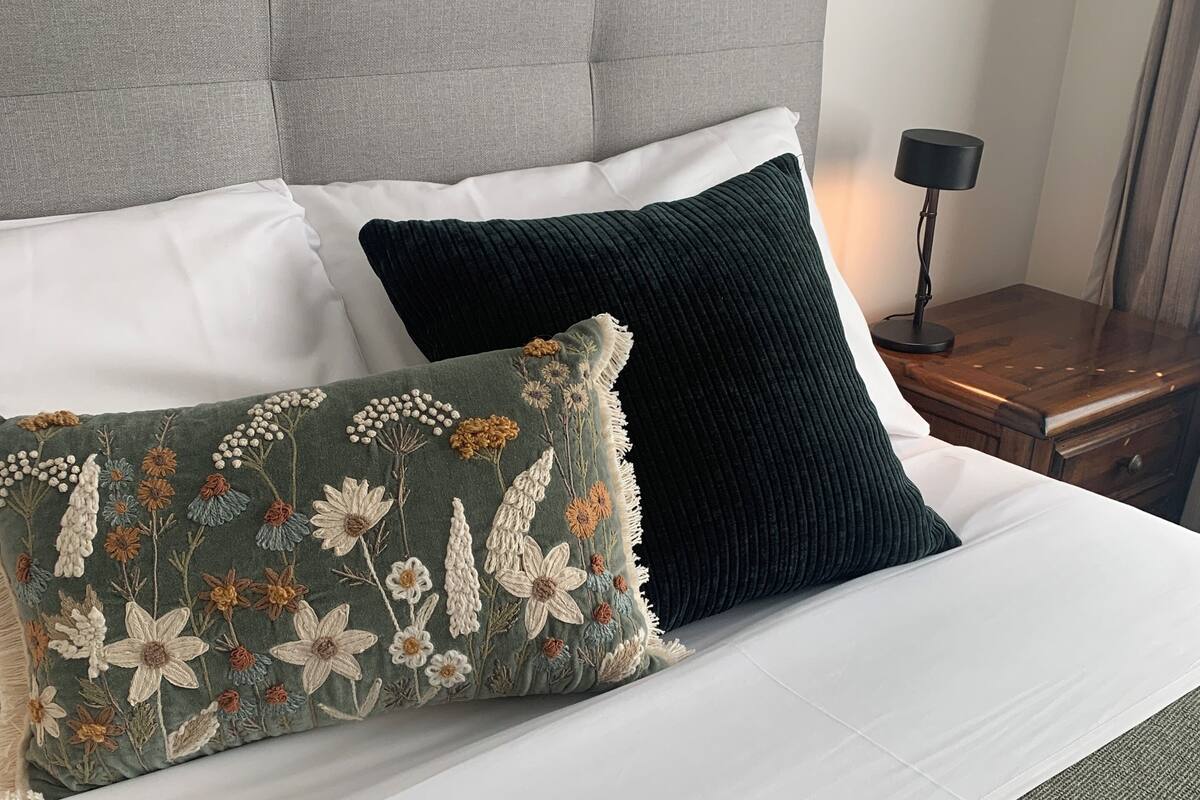 A neatly made bed with a grey upholstered headboard, white bedding, and two decorative pillows with floral patterns.