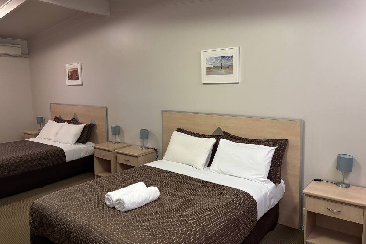 A hotel room with two beds, each with a headboard, bedside tables with lamps, and a framed picture on the wall.