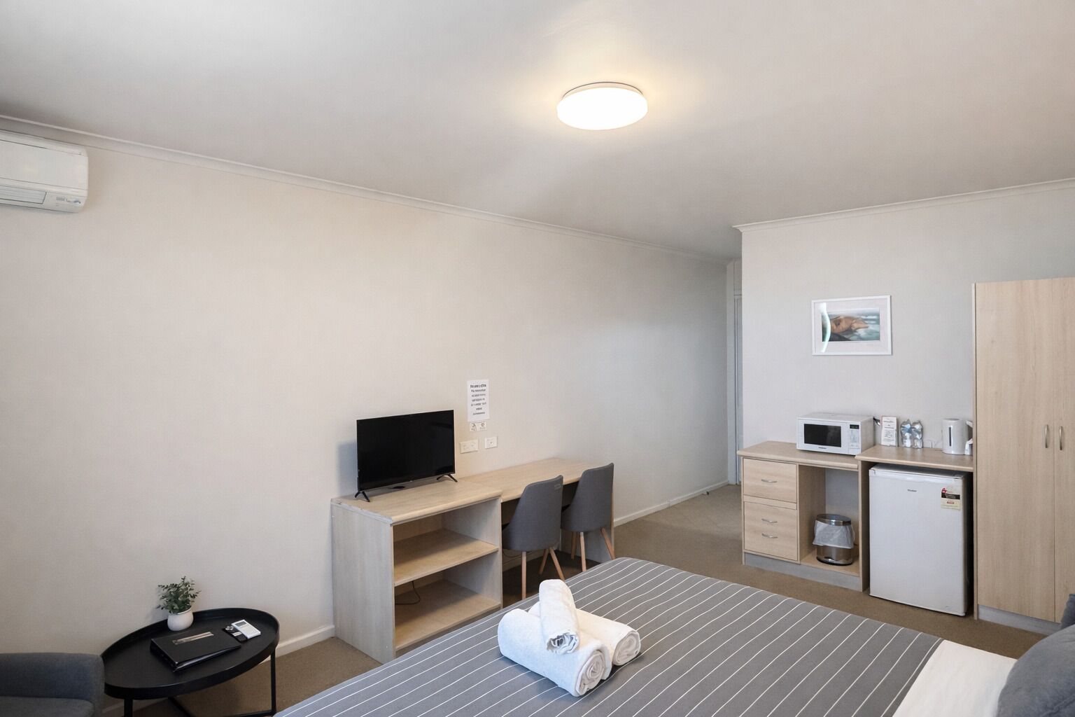 A compact hotel room with a bed, a desk with a TV, a kitchenette, and a small bathroom.