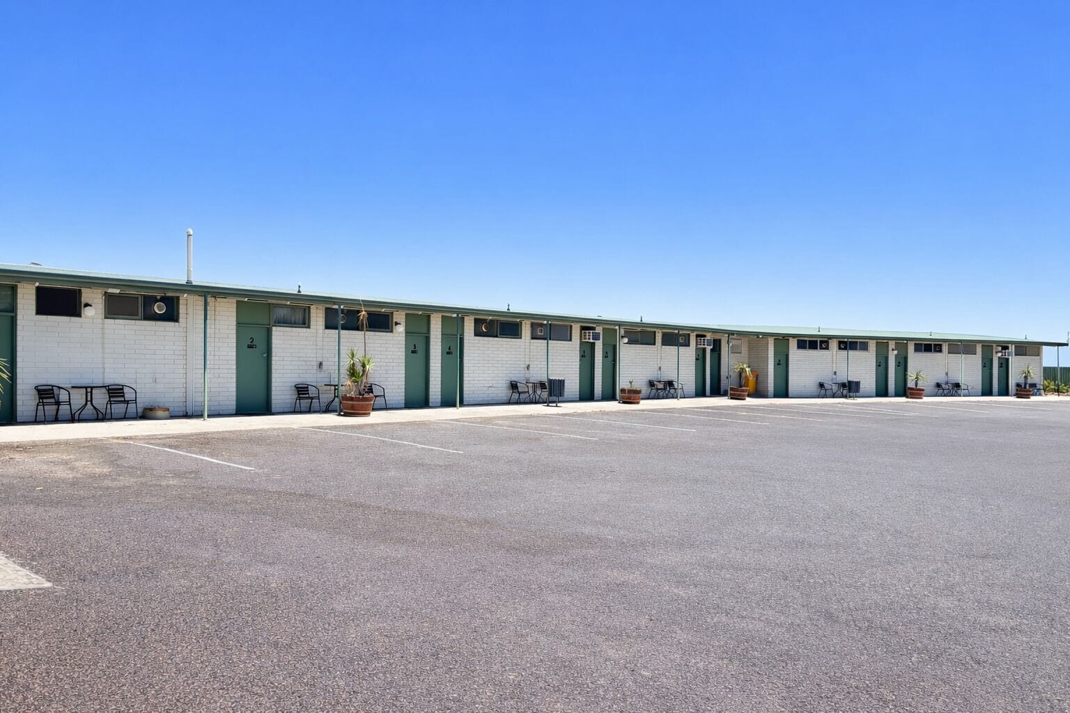 A row of motel rooms with individual doors, a parking lot, and outdoor seating areas.