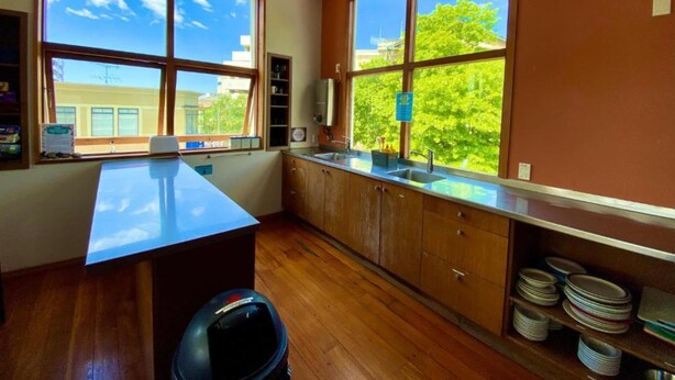 A kitchen with wooden cabinets, a countertop, and a large window offering a view of trees and buildings. - On Top Apartments & Hostel Backpackers (Dunedin)