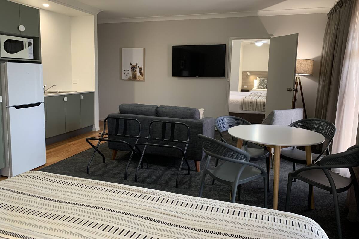 A modern hotel room with a bed, a sofa, a dining table, and a kitchen area with a microwave and refrigerator.