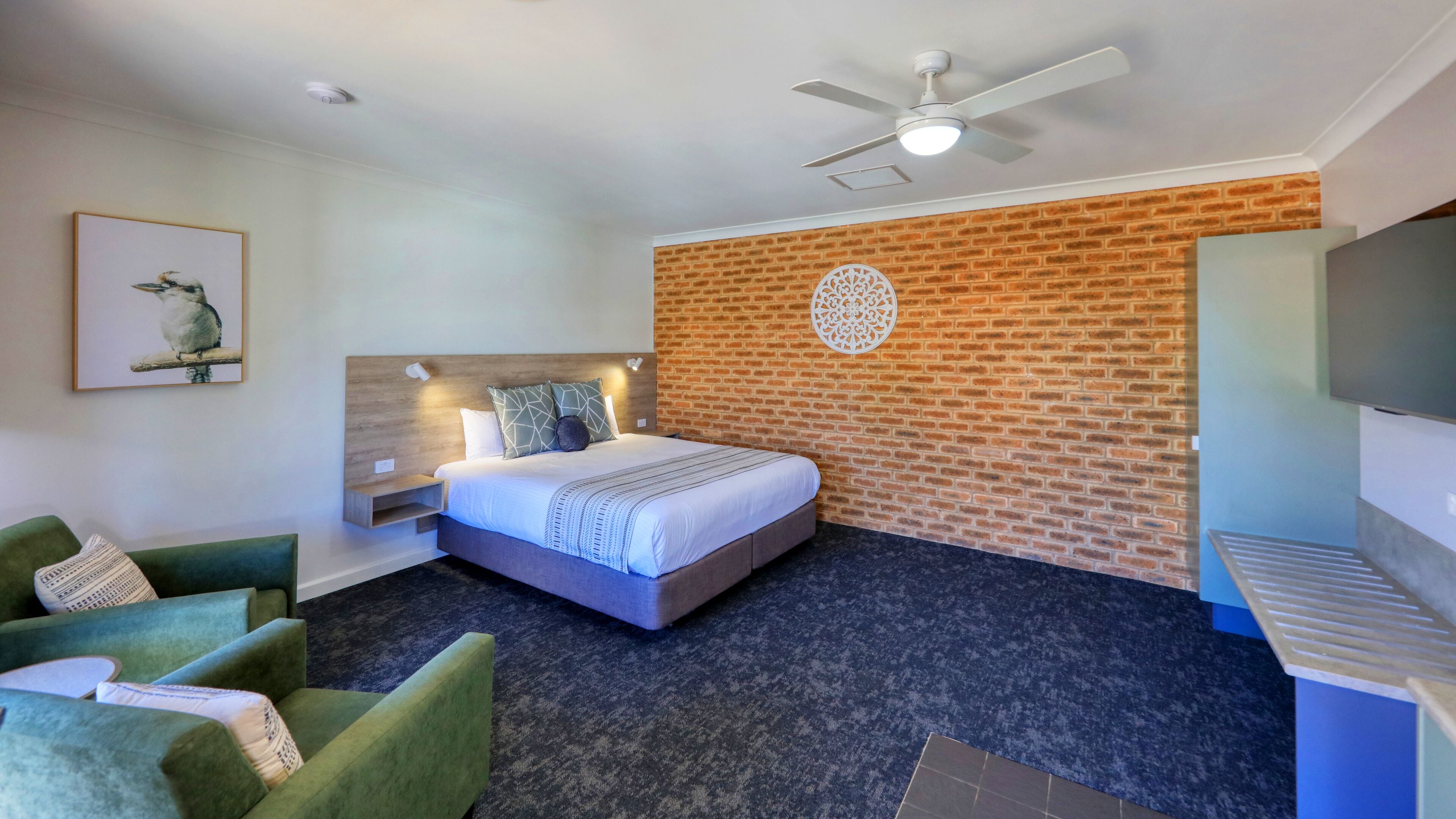 A hotel room with a bed, green armchairs, a brick wall, a ceiling fan, and a TV.