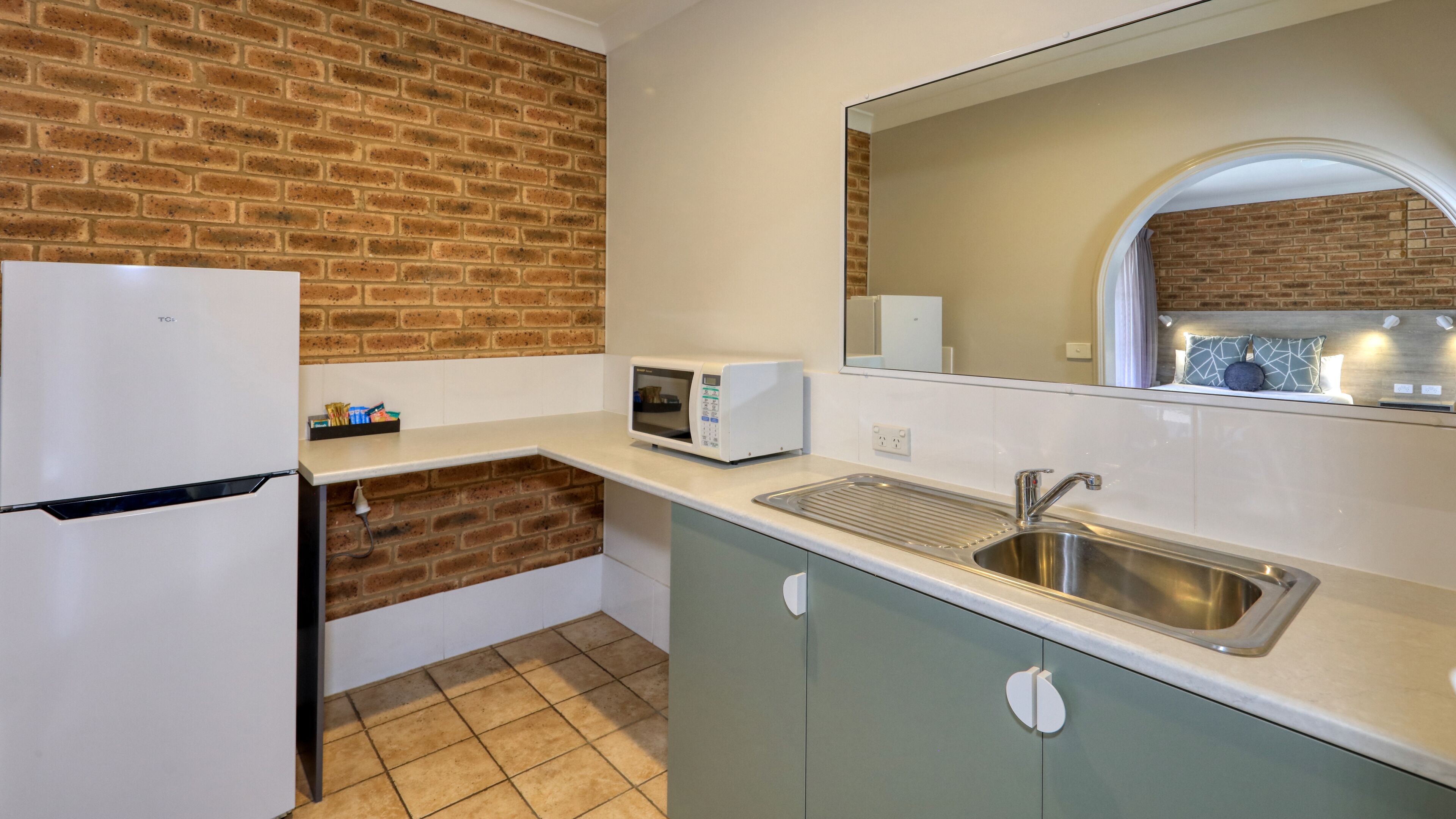 A compact kitchen with a refrigerator, microwave, sink, and a brick wall.