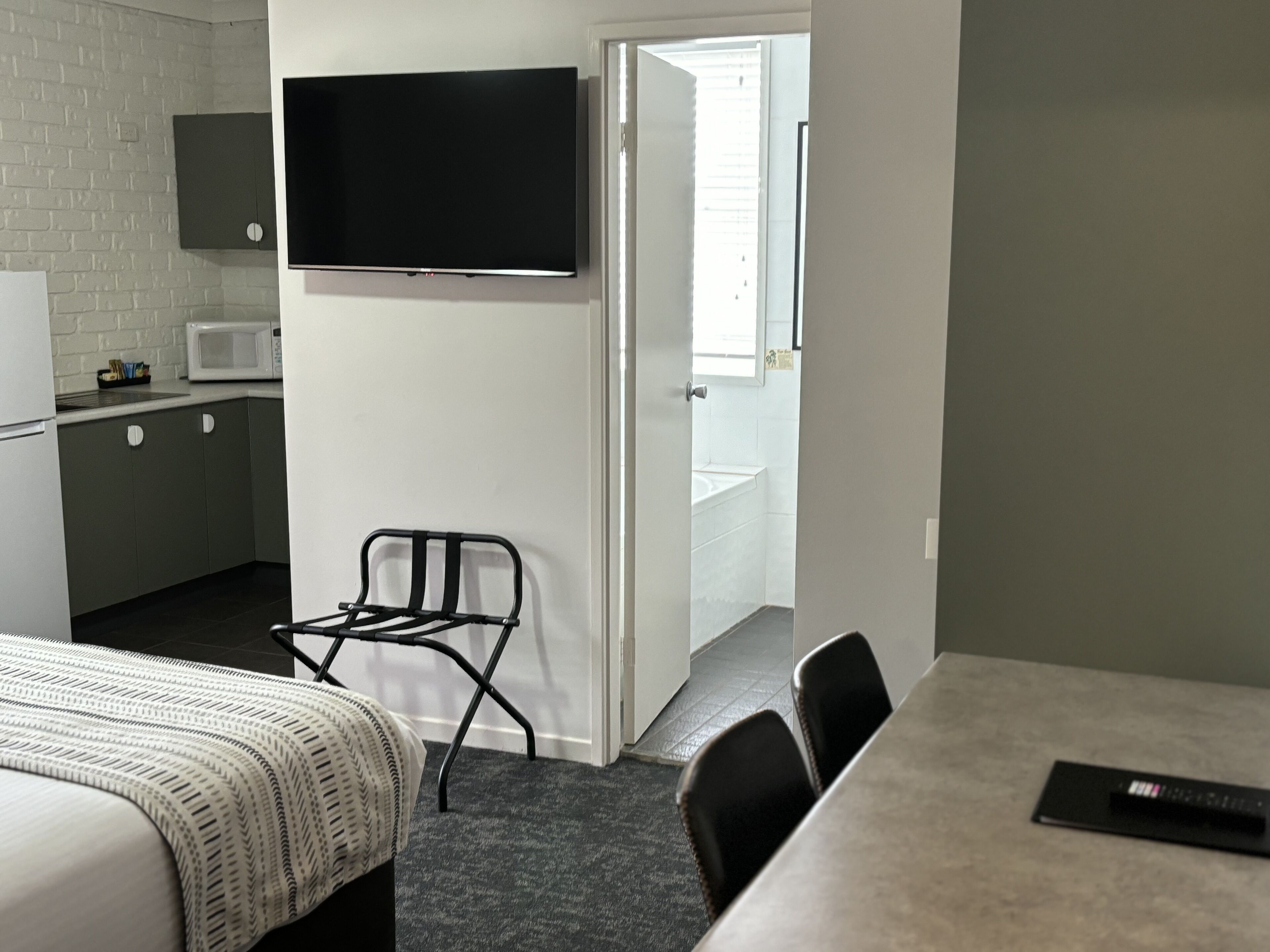 A compact hotel room with a kitchenette, a flat-screen TV mounted on the wall, a dining table with chairs, and a small chair.