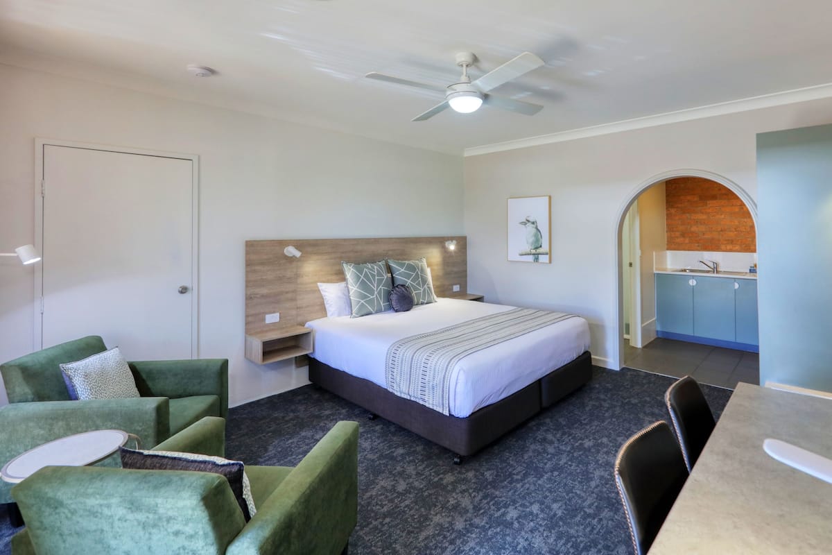 A modern hotel room with a bed, green armchairs, a desk, and a kitchenette.