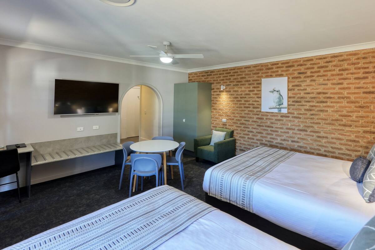 A hotel room with two beds, a desk, a chair, a TV, and a brick wall.