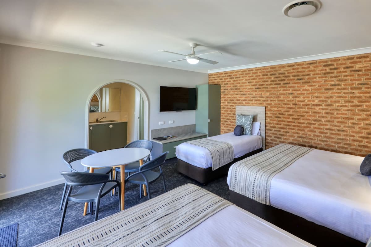 A hotel room with two single beds, a small kitchenette, a dining area with a table and chairs, and a brick wall.
