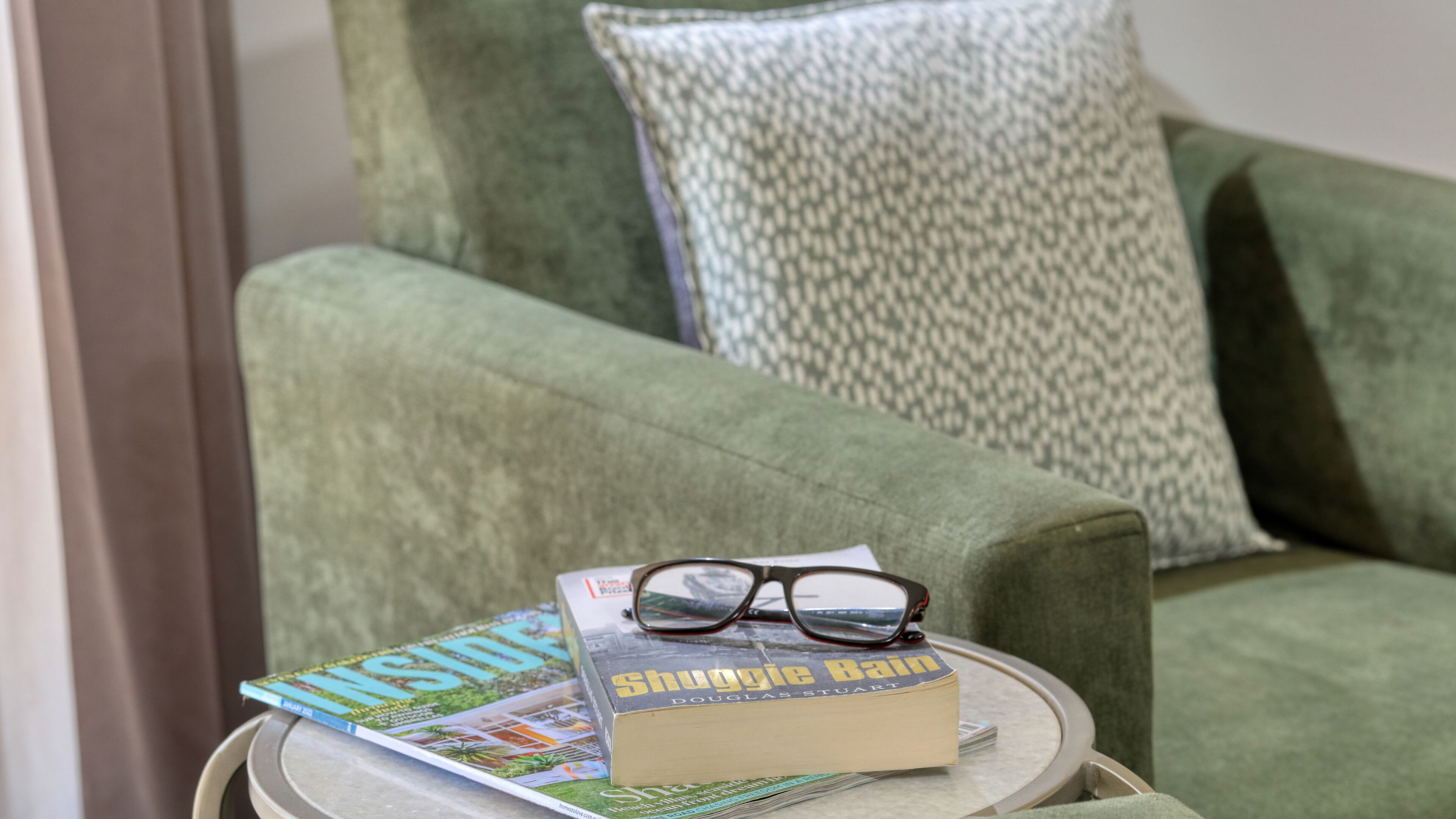 Ein grüner Sessel mit einem gemusterten Kissen, ein kleiner runder Tisch mit einer Zeitschrift, einem Buch und einer Brille.