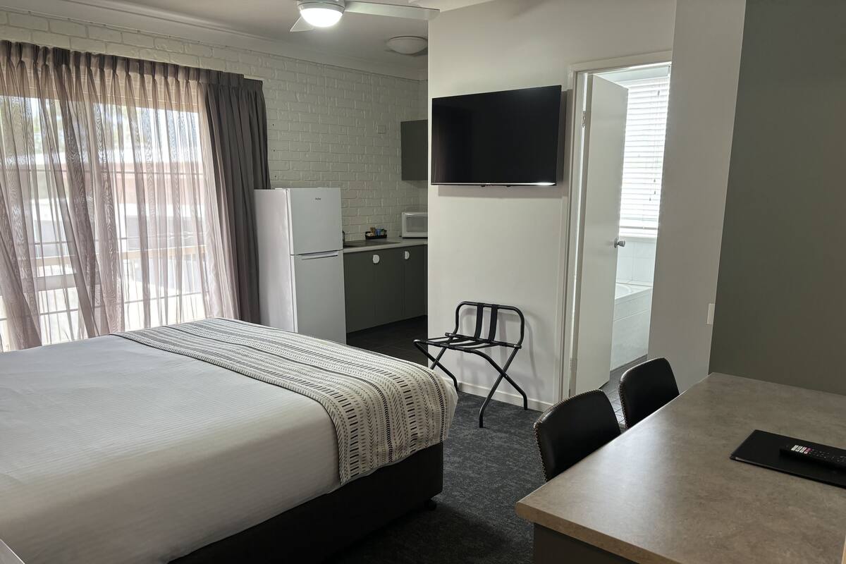 A hotel room with a bed, a desk, a chair, a TV, a kitchenette, and a door.