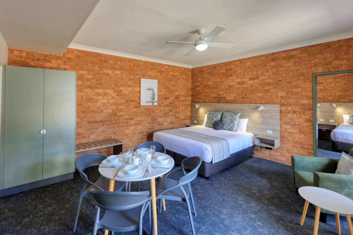 A hotel room with a brick wall, a bed with a wooden headboard, a dining table with chairs, a small table with a chair, and a green armchair.