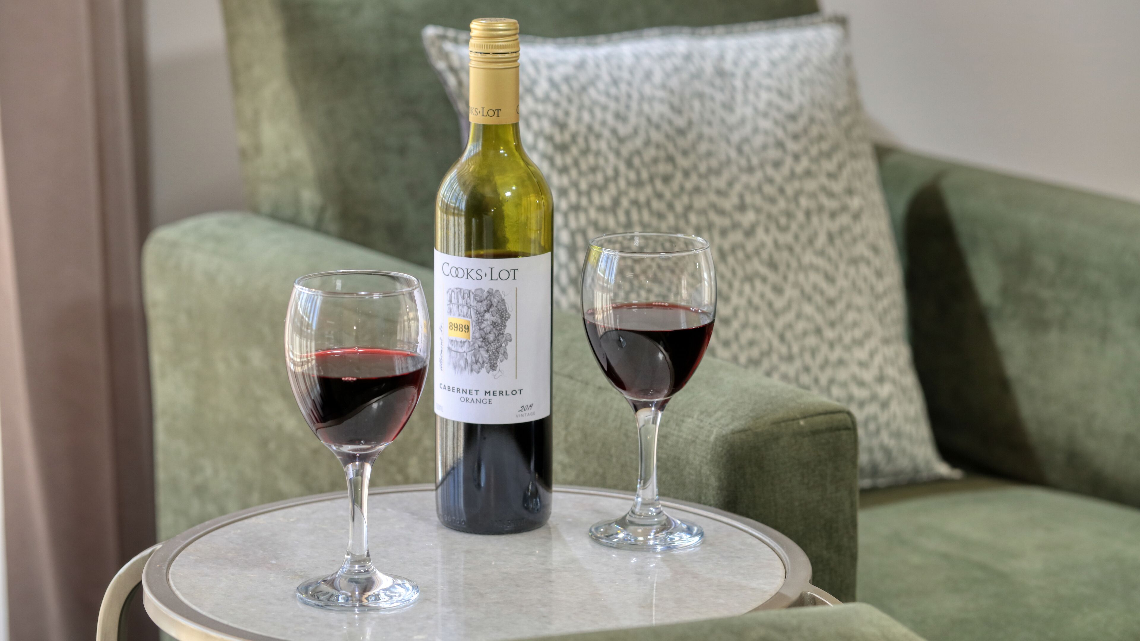 Una botella de Cooks Lot Cabernet Merlot Naranja sobre una mesa redonda con dos copas de vino llenas de vino tinto, colocadas sobre una silla tapizada de verde con un cojín estampado.