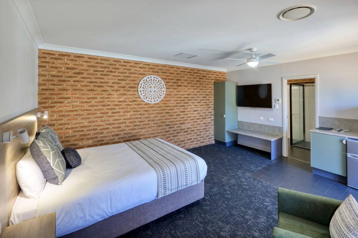 A hotel room with a brick accent wall, a bed with pillows, a green armchair, a small desk with a TV, and a shower area.